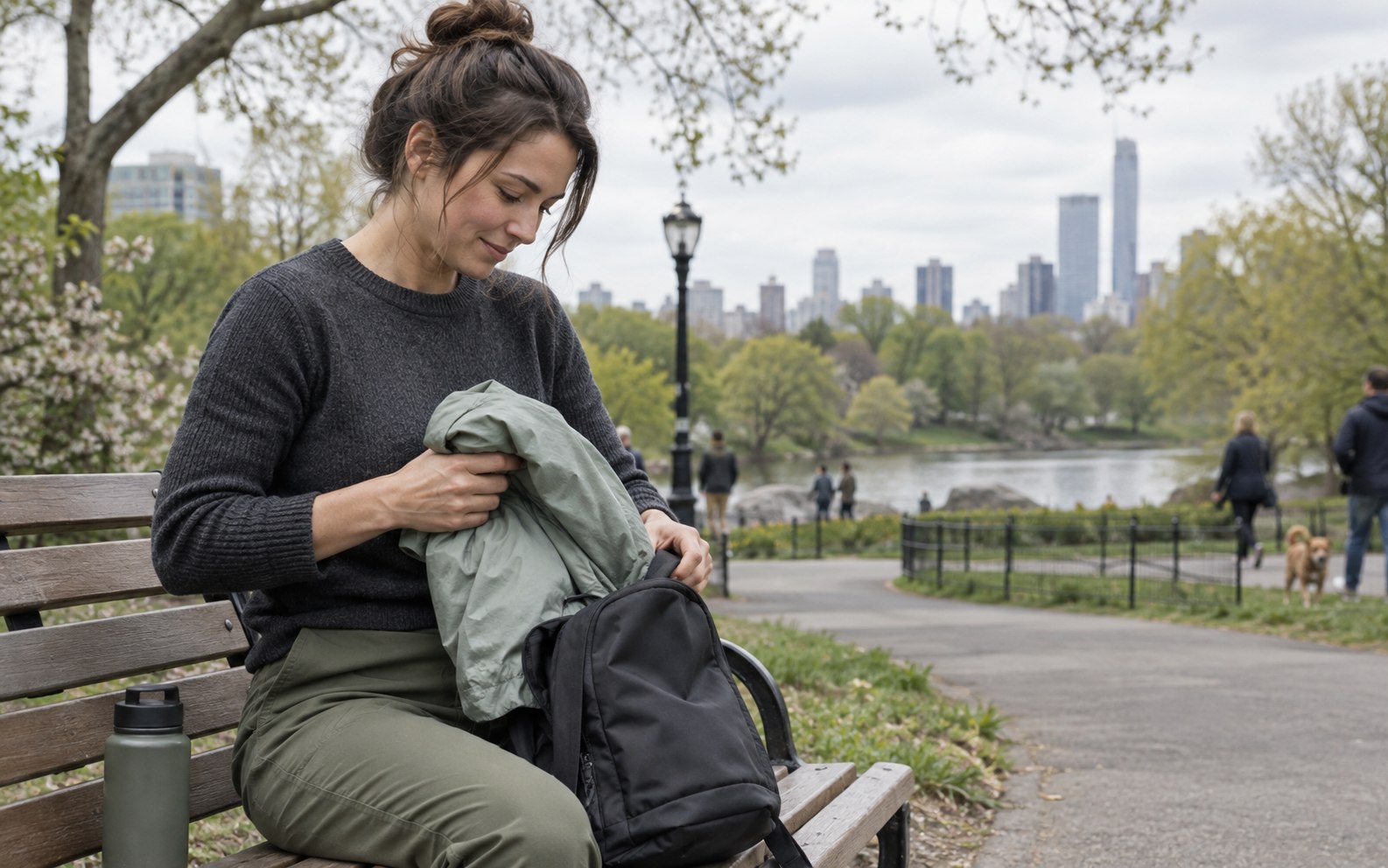 A person packing a lightweight jacket into a day bag beside a city park